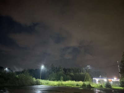 The image depicts a night scene at a gas station on the outskirts of Благовещенка Кп, Russia. A wet, asphalted area, likely a road or parking lot, is visible in the foreground with reflections of lights. Beyond this, a strip of tall, green grass and bushes separates the foreground from a line of trees. In the background, a gas station structure with illuminated pumps and a canopy is visible to the right. Several streetlights are present, illuminating the area with a bright, white light. The sky is dark and overcast with clouds, and there are two small, bright lights visible in the upper right portion of the sky, possibly aircraft or distant lights. The overall atmosphere is dim and illuminated by artificial lights.