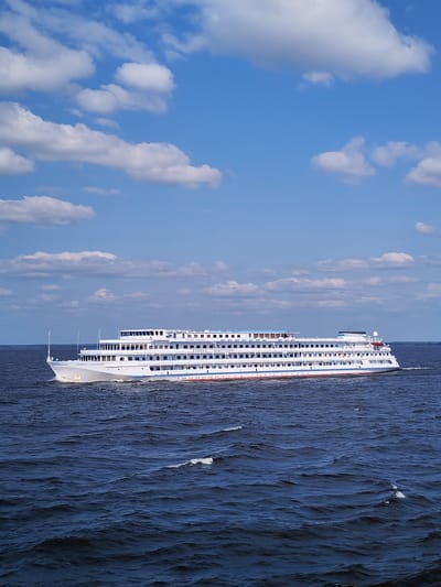 A wide, white, multi-deck river cruise ship is sailing on dark blue water under a bright blue sky with scattered white cumulus clouds. The ship has many windows along its sides and a blue accent along the top decks. The name "КНЯЖНА АНАСТАСИЯ" is visible on the side of the ship. The water is choppy with visible whitecaps. The horizon in the distance is a faint, dark line suggesting land. The image is likely taken from another boat or from the shore looking out onto the water. The scene depicts a sunny day on a waterway, possibly a river or lake.