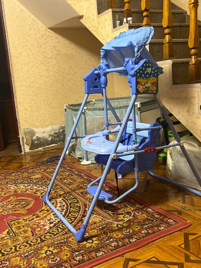 The image depicts an indoor scene, likely a residential setting, with a focus on a blue baby swing set positioned on a rug. The swing set is the primary subject, with its frame, seat, and canopy visible. Behind the swing, a crib or playpen is partially visible. The environment includes a carpet with a red and yellow intricate pattern, a wooden floor with geometric inlays, and a staircase with wooden railings and steps in the background. The walls are textured and painted a light yellow. There are some bottles on the stairs, possibly medicine or cleaning supplies. The lighting suggests an indoor environment, possibly in the late afternoon or evening. The overall impression is of a domestic space with children's items. The location is specified as Jrarbi, Armenia.