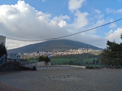 This wide-angle outdoor photograph captures a scenic landscape featuring a prominent mountain, a town, and a paved foreground. The sky above is a mix of bright blue patches and white, fluffy clouds, with some larger clouds partially obscuring the top of the mountain.
Dominating the background is a large, verdant mountain, its slopes densely covered with trees and green foliage, giving it a lush appearance. The upper portion of the mountain is shrouded in a soft layer of clouds, adding depth and atmosphere to the scene. Nestled on the lower slopes and foothills of this mountain is a sprawling town or large village, characterized by numerous light-colored buildings that appear to be made of stone or stucco, contrasting with the green landscape.
In the mid-ground, a vibrant green field or cultivated land stretches between the foreground and the mountain-side town. A tall, dark utility pole with a streetlamp fixture stands somewhat centrally in the mid-ground, interrupting the view of the green field. To the left of this pole, a white flagpole proudly displays the flag of Israel, featuring a blue Star of David and blue stripes on a white background. This area also includes a low white wall or structure.
The immediate foreground is a large, open, paved area. The paving stones are light and dark grey, laid out in an intricate circular or spiral pattern that emanates from the center. There are some darker, irregular patches on the paving, suggesting dampness or wear.
On the far left of the image, part of an off-white building or wall is visible, with dark green foliage growing beside it. Below this, a set of blue metal railings, possibly part of stairs or a viewing platform, descends downwards, supported by a base of dark, rough-hewn rocks. On the far right, a large, dark green tree with dense foliage frames the edge of the scene.
Two dark, thin power lines or communication cables stretch horizontally across the upper middle of the frame, passing in front of the clouds and the mountain.
The lighting suggests it is daytime, possibly late morning or early afternoon, with a mix of sun and clouds. The overall impression is one of a peaceful, well-established settlement set against a grand natural backdrop.