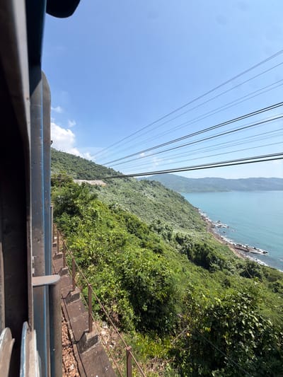 The image shows a view from what appears to be a moving tram or cable car, likely in the Hải Vân Ward, Vietnam, given the lush green mountainous terrain meeting the ocean. Several parallel overhead cables stretch across the frame diagonally, disappearing into the distance. To the right, dense green foliage covers the hillside leading down to a rocky coastline and the ocean. The sky is a pale blue with a few scattered clouds. The bottom portion of the image shows the interior of the tram or cable car, with metal railings and a slightly obscured view of the landscape. An old concrete staircase and rusted metal rail is to the bottom right, below the train car.
