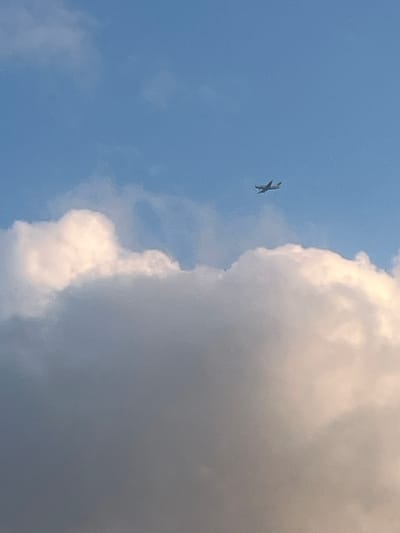 The image shows a small, propeller-driven airplane flying in a blue sky above clouds. The airplane is silhouetted against the sky and appears to be a light aircraft. The clouds are white and fluffy, with some areas appearing darker and more shadowed, suggesting they are large cumulus clouds. The lighting indicates it is daytime, possibly late afternoon or early morning, as the clouds have warm tones. The image does not contain any specific landmarks or details that would indicate a location within Jacksonville, United States of America. There are no people or interactions visible in the image.