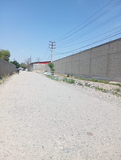 The image depicts a desolate dirt road in Eshanguzar, Uzbekistan. The road surface is composed of packed earth and gravel. On either side are high, unfinished gray brick walls. Litter and debris are scattered along the base of the walls. Power lines stretch across the sky above the road. In the distance, a person walks along the road. Several vehicles are also visible further down the road. The sky is clear and blue. A utility pole stands on the right side of the road.