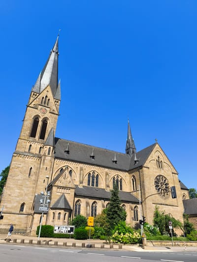 The image shows the St. Johann Church in Georgsmarienhütte, Germany, under a clear blue sky. The church is constructed of light brown stone and features a tall, pointed spire with a clock face. The architecture is Gothic, with pointed arch windows and a large rose window on the facade. The church is surrounded by green trees and bushes, with a road in the foreground. A traffic light is visible on the right side of the image. A person wearing a blue jacket and shorts is walking on the sidewalk to the left of the church. A street sign with a train station symbol and the word "Bahnhof" is posted on a pole near the church entrance. Another yellow sign with "U2" and an arrow is visible to the right of the church.