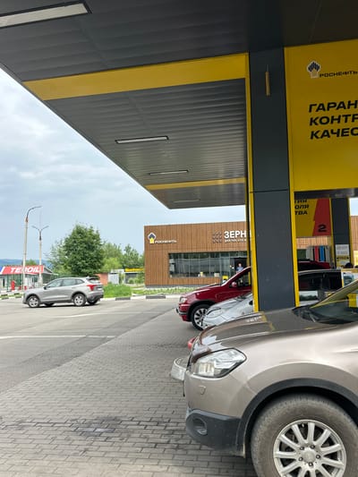 The image shows a gas station canopy with a yellow and gray design. Several cars are present, including a silver SUV, a red SUV, and a tan SUV in the foreground. The gas station has signage that appears to be in Russian, with the logo "РОСНЕФТЬ" (Rosneft) visible. There are also advertisements and numbers on the yellow pillars. In the background, there are buildings, trees, and a cloudy sky. One building has the text "ЗЕРН" (likely "Zerno" meaning "grain") and some additional Russian text. Another structure has "TETOIL" written on it. The scene depicts an ordinary day at a gas station in Slyudyanka, Russia.