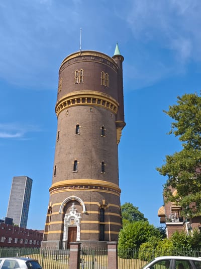A water tower, identified as the Watertoren West, stands tall in the city of Tilburg, Netherlands. The tower, constructed of dark brown brick with yellow horizontal bands and decorative yellow trim, features numerous arched windows. A conical green spire crowns a section of the tower. In the background, a modern, tall, grey skyscraper with a grid-like facade is visible, along with lower, red-brick buildings. Lush green trees and bushes surround the base of the tower and other structures. The sky is a clear, bright blue. A metal fence runs along the foreground, and the tops of two cars are visible behind it. No people are present in the image.