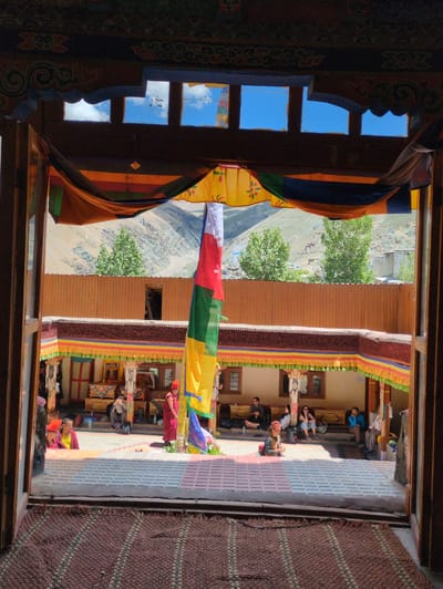 The image depicts a courtyard of a Tibetan monastery, possibly in Pibiting, India, viewed through an open wooden doorway. The foreground features a woven rug. The courtyard itself is paved with square tiles. People are seated and standing around the perimeter of the courtyard. In the center of the courtyard, a tall, multi-colored prayer flag hangs vertically, extending from the upper structures down to the ground. A monk wearing a maroon robe and an orange hat stands near the flag. Other individuals in the courtyard are dressed in casual clothing, some appearing to be tourists. The background reveals a building with a corrugated metal wall and mountains beyond, under a bright blue sky with some clouds visible through square openings in the overhead structure. Colorful prayer banners are draped along the covered walkways of the courtyard.