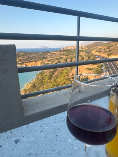 A close-up shot focuses on a glass of red wine positioned on a speckled grey surface. Behind the glass, out of focus, a second glass filled with an orange beverage is visible. The background reveals a scenic view of the sea and coastline, with a road winding through a hilly, vegetated landscape. The sea is a clear turquoise, meeting a sandy beach and rugged cliffs. In the distance, a small island or prominent landmass is visible on the horizon under a clear blue sky. The overall scene suggests a relaxed setting, likely a balcony or patio overlooking a coastal area in Agios Nikolaos, Greece.