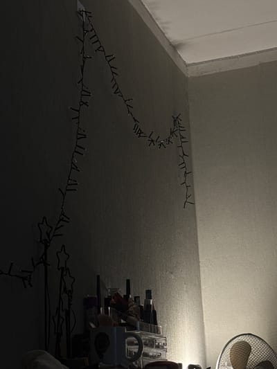 The image depicts an interior scene, likely a bedroom or living space, in a residence located in Olaine, Latvia. The focal point is a string of fairy lights draped across a dark gray wall, with some lights appearing to be attached by small clear clips. The lights are unlit and form a somewhat haphazard trail, starting from the upper left and curving down and to the right. To the left, partially visible, are decorative star-shaped wire frames, also appearing to be part of a light display or decoration. In the lower portion of the image, a collection of objects is arranged on a surface, including a white mug with a design, a clear plastic organizer filled with various items (possibly cosmetics or art supplies), and a black pen. To the right, a desk fan with a metal grille is partially visible, illuminated by an unseen light source, casting a warm glow on the adjacent wall and ceiling. The overall ambiance is dim, with shadows dominating the left side of the frame and a brighter, warmer light on the right. No people are present in the scene.