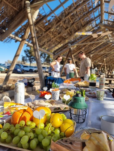 In the city of Palmachim, Israel, a group of people are gathered under a thatched roof structure, likely for a picnic or outdoor meal. The scene is set on a sandy surface, with a table laden with food and drinks. In the foreground, a spread of fresh fruits is visible, including green grapes, sliced oranges, and yellow tomatoes. Wrapped sandwiches or wraps are also present, along with containers of cut vegetables like carrots and celery sticks. A green lantern sits on the table, adding to the outdoor ambiance. Beverages include a can of what appears to be beer and a bottle of infused water with mint and lemon. In the background, several cars are parked, suggesting a day trip or outing. Three individuals are partially visible, engaged in conversation. The overall atmosphere suggests a relaxed and social gathering in a coastal or beachside setting, characteristic of the Palmachim area.