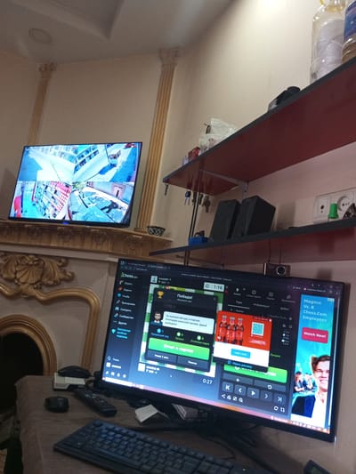 The image depicts an indoor scene with multiple electronic devices and furniture. In the foreground, a computer monitor displays a chess website interface, showing game statistics, chat messages, and an advertisement. A keyboard and two computer mice are positioned on a table in front of the monitor. To the left of the monitor, a large flat-screen television shows a multi-camera security feed of what appears to be a store or public space. The background includes a fireplace with decorative elements, and shelves holding various items, including speakers and bottles. The overall impression is of a domestic or office setting where entertainment and surveillance systems are in use. The location of Kattakurgan City, Uzbekistan, is not visually identifiable from the provided image.