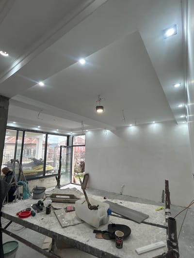 The image depicts the interior of a room undergoing renovation in Dolni Tsibar, Bulgaria. The room features freshly plastered, white walls and a multi-tiered white ceiling with numerous active recessed LED spotlights and one central suspended cylindrical light fixture. Exposed electrical wires hang from the ceiling at various points.

On the left, large glass windows and a partially open glass door, set in dark frames, provide a view to the outside. Visible outdoors are a bright yellow personal watercraft (jet ski) parked on a stand or trailer, residential buildings with tiled roofs, and bare trees or shrubs, suggesting an overcast day.

In the foreground and midground, a temporary work platform or scaffolding is present, heavily dusted with white plaster or drywall compound. Scattered on this platform are various construction tools: a white rectangular plaster float marked "САЛИ" (SALI), several smaller hand trowels and spatulas, a red plastic container, a black coffee cup with a logo, and a large translucent plastic jug containing a mixing tool. A single gray rectangular tile or board is also on the platform, with another similar tile lying on the concrete floor near the right wall. Metal scaffolding poles support the platform on the far right.

To the far left, partially obscured, an adult male with light hair is seated on a step ladder, facing away from the camera. He appears to be a construction worker, possibly engaged in detail work on the lower wall or preparing materials. Another step ladder and two buckets (one dark, one lighter green) are visible near him, adjacent to the exterior window. The concrete floor throughout the room is covered in dust and debris, consistent with active construction work.