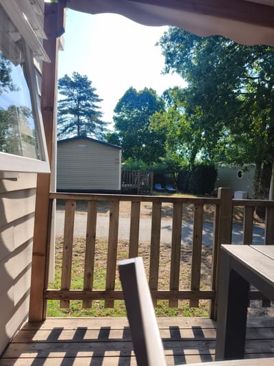 The image depicts a view from a wooden porch, looking out towards a sunny outdoor scene. In the foreground, a portion of a light-colored mobile home or caravan is visible on the left, with its siding and an open window. The porch itself is made of wooden planks, and a wooden railing with vertical posts and horizontal supports runs across the mid-ground. Beyond the railing, a gravel path is visible, lined with grass and fallen leaves.
Further into the scene, several trees with green foliage dominate the background, with a bright, clear sky above. Two light-colored reclining chairs are situated on the grass in front of a hedge. Two mobile homes or caravans are visible in the distance. One, closer to the left, is light beige with a dark roof and the text "O'HARA" and the number "404" on its facade. Another caravan, to the right, is light green and has a circular window.
The image appears to be taken in a campground or a residential area with mobile homes, likely during daytime, given the bright sunlight casting distinct shadows on the porch. The location is identified as Pierrefitte-sur-Sauldre, France. There are no people visible in the image.