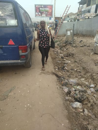A woman wearing a floral blouse, black pants, and a white headscarf is walking on a dirt road in the city of Way, Nigeria. The road is dusty and there is a pile of trash on the side of the road. A blue van is parked on the side of the road. There is a sign in the background with an image of a man in a suit. The sign advertises an event called "Wake n' Praise." There is a ladder leaning against a building in the background.