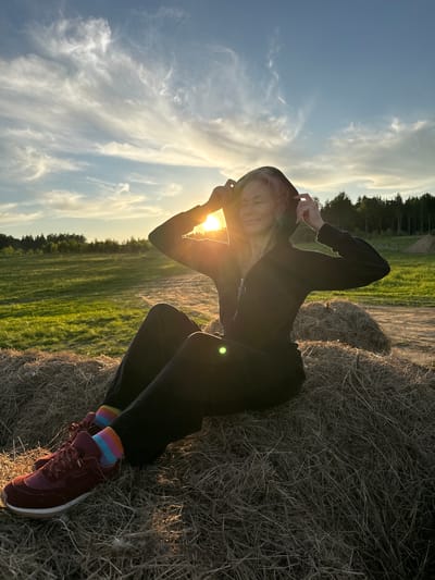 The image depicts a young individual, appearing female, seated or leaning on a large hay bale in an open, rural landscape, likely in Стеклянный, Russia. The person has light-colored hair and is wearing a dark hooded sweatshirt or jacket, dark pants, colorful striped socks, and maroon sneakers. Their hands are raised near their head, somewhat shielding their face from the sun, which is low on the horizon and creating a strong backlighting effect with prominent lens flare.

The scene captures the golden hour of sunset, indicated by the warm, golden light illuminating the person and parts of the environment. Behind the subject, a vibrant green field stretches towards a line of dark trees in the distance. The sky above is a blend of clear blue and scattered, wispy clouds, softly lit by the setting sun. The hay bale provides a natural seat, its dry texture visible in the light. The individual's expression, though partially obscured, suggests a sense of contentment or a gentle smile, as if enjoying the moment outdoors.

The activity is posing for a photograph, possibly while relaxing in the evening light. There is no visible text in the image. The original image is rotated 90 degrees counter-clockwise, causing the horizon to appear vertical and the person to be oriented diagonally across the frame.
