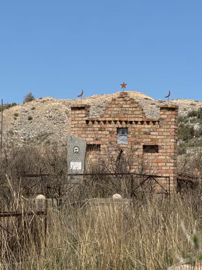 The image shows a brick structure with a weathered appearance, possibly a mausoleum or memorial, in what appears to be a dry, grassy field. The structure has a star and crescent shape attached to it. There is a fence and some grave markers in front of the structure. The location may be Barbulak, Kyrgyzstan. The sky is clear and blue. The image appears to be taken during the day.