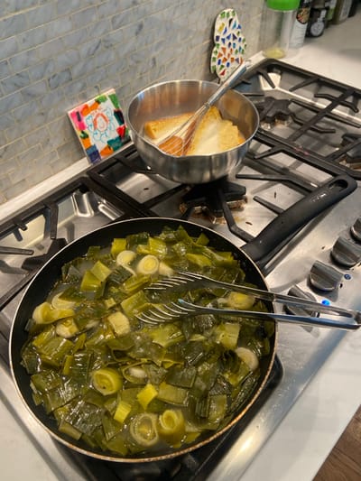 A close-up image of a kitchen counter in Rye, New York, taken in a residential setting. The scene is a kitchen counter with a stovetop. The stovetop is stainless steel and has two burners. The left burner is on and has a black skillet containing cooked leeks. The leeks are green and yellow and are in a clear liquid. A pair of metal tongs is resting on the skillet. The right burner is off and has a stainless steel pot with a white substance in it, likely a custard. There is a whisk in the pot. There is a small piece of artwork behind the pot, which is a hand-drawn picture on a ceramic tile. The scene is lit by overhead lighting. The image appears to have been captured in a residential kitchen, with a typical kitchen counter surface. The scene is of a cooked meal, likely prepared for consumption.