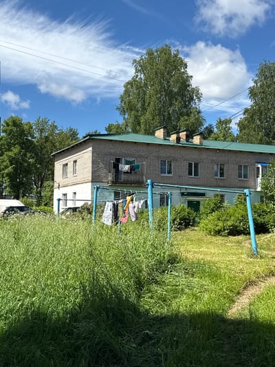 The image depicts a scene in Valday, Russia, on a sunny day with a blue sky and wispy clouds. In the foreground, a field of tall, green grass dominates the lower portion of the frame. A blue clothesline, supported by two blue posts, is set up in the grass, with several items of clothing hanging on it, including shirts and pants. Behind the clothesline, a two-story brick building with a green roof is visible. The building has multiple windows and a balcony on the second floor where more laundry is hanging. The building appears to be residential. Trees with lush green foliage surround the building, and a white van is partially visible to the left of the building. The overall impression is of a rural or suburban setting.