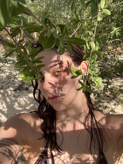 A female individual is photographed in a close-up, chest-up shot on a sandy beach in Baan Nai Wok, Thailand. Her dark, wet hair falls around her shoulders. Her face features fair skin, amber/hazel eyes looking directly into the camera, and slightly parted lips. Green foliage from a bush or tree is positioned in the immediate foreground, partially obscuring the upper face and body, and casting distinct shadows across her skin. She is wearing a dark, possibly patterned, bikini top. A tattoo with dark horizontal lines is visible on her left upper arm. A subtle red mark is present on the upper right side of her chest, near the clavicle. The background consists of light-colored sand and additional green vegetation under direct, bright sunlight.