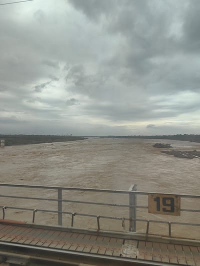 The image shows a wide, muddy river flowing under a cloudy sky. The river has strong currents and appears to be swollen, suggesting recent rainfall or a flood. On the right side of the riverbank, there are some rocky outcrops. In the foreground, there is a metal railing and a wooden walkway, indicating a bridge or a pier. A yellow sign with the number "19" in black is attached to the railing. The background shows a densely vegetated riverbank with some distant structures. The overall scene depicts a natural environment affected by water, under an overcast and potentially stormy sky. The location is identified as Kandari, India, which suggests a specific geographical context for the river's conditions.