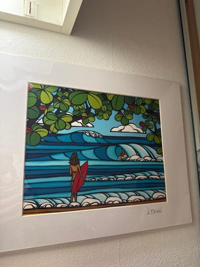 A framed picture is hanging on a white wall. The picture depicts an ocean scene with a woman standing on the beach, looking out at a wave. She is holding a red surfboard. There are other waves in the background, and a smaller person is surfing one of the waves. The picture is signed "H. BROWN" in the bottom right corner. The scene is depicted in a cartoonish style. The picture is hanging on a white wall in a room in a house in Takarazuka, Japan, a city located at latitude: 34.811, longitude: 135.342.