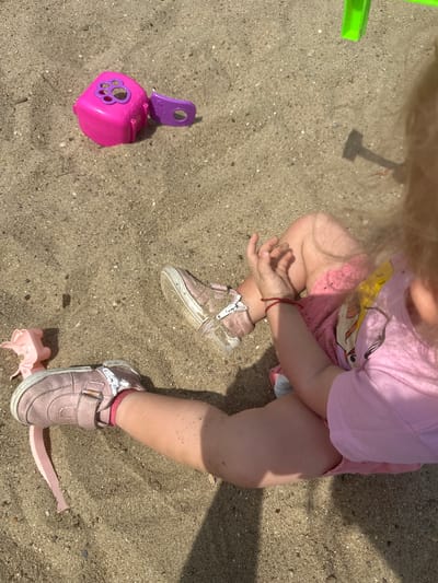 The image shows a child sitting on a sandy surface, likely a playground or beach. The child is wearing pink shoes and a pink outfit. Scattered around the child are various toys, including a pink sand toy with a paw print design and a purple attachment, and a pink plastic piglet. A green structure, possibly part of a playground, is visible in the upper right corner. The ground is covered in sand, with some small stones and debris. The lighting suggests it is daytime, with shadows cast on the sand. The location of Donchovci, Bulgaria is not discernible from the image itself.