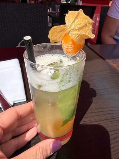 A cocktail glass with a black straw sits on a dark brown wooden surface. A golden husk fruit garnish adorns the rim. The glass contains a frothy, light-colored drink, with visible green leaves. A hand with a purple fingernail holds the glass. A white folded napkin lies underneath. An arm and part of a red chair are visible in the background. This image was likely taken at a cafe or bar. There is no information that can determine this image was taken in Guildford, United Kingdom.