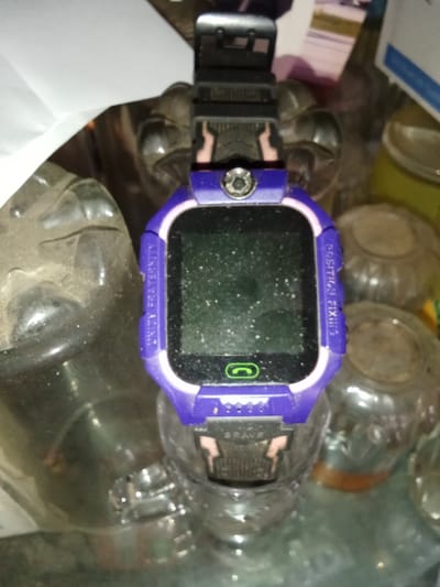 A purple and black children's smartwatch, model type Q12, is positioned centrally in the frame, resting on top of an inverted, clear, ribbed glass jar. The smartwatch features a square, dark, and heavily dust-speckled screen. A small, circular silver camera lens or decorative element secured by a screw is located above the screen. The screen's lower bezel displays a green telephone receiver icon. The left side of the watch body bears the inscription "UNITY FRATERNITY," and the right side reads "POSITION FIXING." Below the screen, the word "BRAVE" is embossed. The watch strap is black with pinkish-beige "lightning bolt" designs at the lugs.
The immediate surroundings consist of multiple clear glass or plastic jars and bottles, all visibly coated in a layer of dust, suggesting a cluttered and uncleaned environment. To the left, a larger, dark, indistinct object, possibly another container, is partially visible and also dusty. A folded piece of white paper is present in the upper left background. No individuals or specific actions are depicted. The scene is an indoor setting within what appears to be a domestic or storage area, located in Emthanjeni Local Municipality, South Africa.