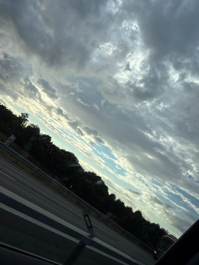 The image shows a diagonal view of a road with trees lining the side. The sky is filled with dramatic clouds, with patches of blue sky visible. The lighting suggests it could be early morning or late afternoon. The scene appears to be a highway in a rural or suburban area. The location is identified as Löftaskog, Sweden, though no specific landmarks or distinguishing features confirm this.