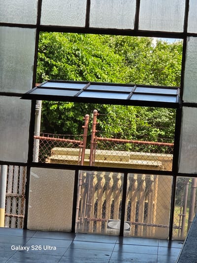 The image shows a view through a multi-paned, frosted glass window with a metal frame. Part of the window is open, revealing a bright outdoor scene. In the foreground, a dark tiled floor is visible. The background consists of lush green foliage, a chain-link fence with barbed wire on top, and rusty metal posts. The sky is visible through the trees. A watermark at the bottom left corner of the image reads "Galaxy S26 Ultra". The specific location in West Hartford, United States of America is not identifiable from the image.
