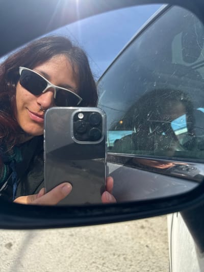 The image displays a selfie taken in the side mirror of a vehicle in Tolhuin, Argentina. A female individual with long, dark hair, wearing silver-framed sunglasses and a nose ring, is reflected, showing a faint smile. Her left hand holds a dark-colored smartphone, visible with a triple camera array and a lidar sensor in a clear protective case, positioned to capture the reflection. Bright sunlight from the upper left illuminates a clear blue sky reflected in the mirror. The adjacent vehicle window displays numerous scratches and minor reflections, possibly of the car's interior. The ground visible below the mirror is a light-toned paved surface.