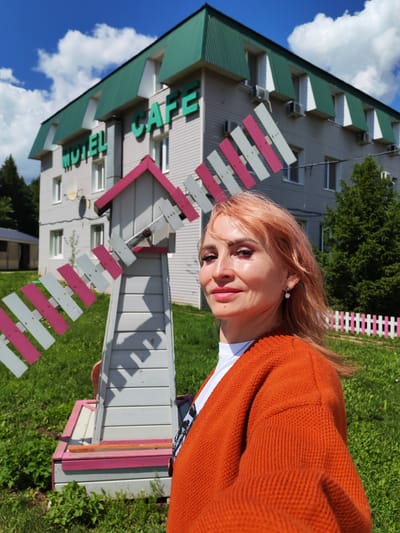 In a sunlit outdoor scene in Baskan, Russia, a woman with peachy-pink hair stands in front of a building labeled "MOTEL CAFE" in green lettering. The building has a light gray exterior with multiple windows and air conditioning units. A decorative windmill with pink and white blades is positioned in front of the building, to the left of the woman. She is wearing an orange knitted cardigan over a white shirt and a silver necklace. The woman is looking towards the camera with a slight smile, her makeup including eyeshadow and mascara. The setting is grassy with trees in the background, and a pink and white picket fence is visible to the right. The sky is blue with scattered white clouds, suggesting a pleasant, daytime weather condition. The overall mood appears relaxed and cheerful, possibly indicating a vacation or a stop at a roadside establishment.