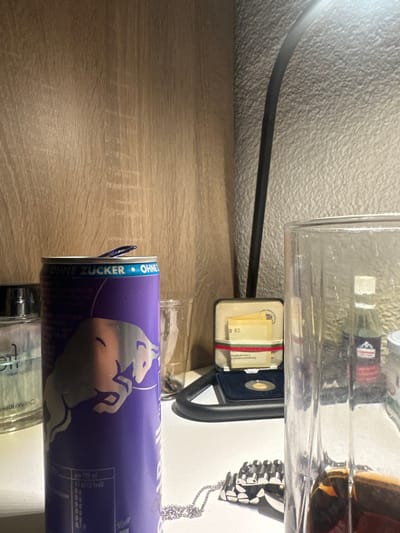 The scene is an indoor setting on a white surface, likely a desk or table, located in Höri, Switzerland. The background consists of a light wood-grain panel on the left and a textured light-grey wall on the right. Illumination suggests a direct light source from above right. No individuals are present.
Objects present include:
* A purple Red Bull "Ohne Zucker" energy drink can, with its tab open, occupies the foreground left. Its branding and nutritional information are partially visible.
* Behind the Red Bull can, a clear glass bottle with a silver-colored cap, partially labeled "Calvin Klein," is visible.
* A clear, empty glass tumbler is positioned between the energy drink can and a desk lamp.
* A black desk lamp with a flexible stem extends vertically from the mid-ground.
* To the right of the lamp, an open cream-colored presentation box contains a gold-colored coin or medallion set against a blue velvet lining. A white card displaying "0 83" and German text, secured by a red and green striped ribbon, is also inside the box.
* In the foreground right, a clear glass pitcher or large tumbler is partially filled with a dark brown liquid and exhibits condensation on its exterior.
* A silver-colored chain necklace with a black and white pendant rests on the white surface below the large glass.
* Reflected within the liquid of the large glass, a miniature clear bottle with a white cap and a red label, partially identifiable as 'Appenzeller', is visible in the background.