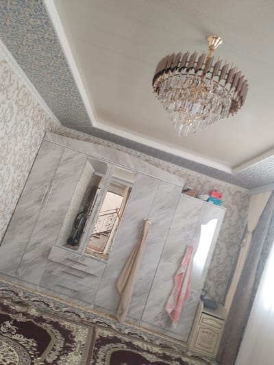 The image shows a bedroom in Kyrgyzstan. The room features a large, marbled wardrobe with mirrored doors. A chandelier with crystal accents hangs from the ceiling, which is decorated with patterned wallpaper and a geometric border. A patterned rug covers the floor. A towel hangs from the wardrobe, and a small chest of drawers is visible on the right side of the room. The scene is indoors, with natural light coming from a window on the right, suggesting it is daytime. The overall impression is of a well-decorated and tidy room. There is no visible text in the image.