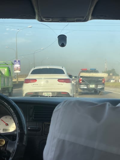 The image shows a view from inside a vehicle, looking out through a cracked windshield. In the foreground, parts of the dashboard and steering wheel are visible. In the midground, there is a white Mercedes-Benz GLE 43, followed by a black pickup truck carrying a mattress. Streetlights are also visible. In the background, the sky appears hazy. A billboard can be seen on the left, below the Mercedes. The license plate of the Mercedes reads "ARC-354MR" indicating that the image was taken in Jiru, Nigeria. The speedometer shows that the vehicle is moving at 20km/h.