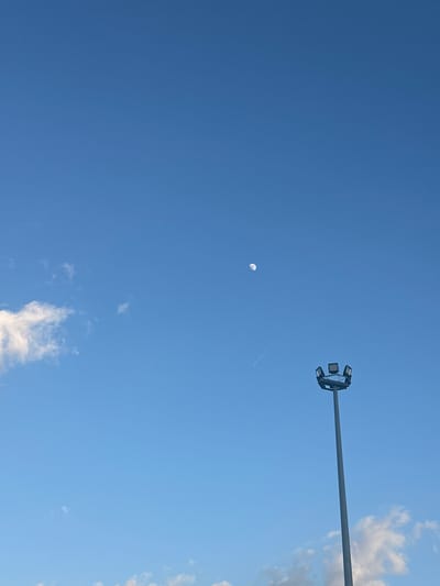 The image depicts a daytime sky in Manisa, Turkey. A light blue sky is visible, with a crescent moon towards the center-left. A light pole with multiple spotlights extends from the upper right corner, pointing towards the center of the frame. Wispy clouds are present along the right edge of the image and toward the bottom. A contrail from an aircraft is visible near the moon.