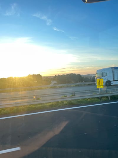 The image depicts a multi-lane highway scene in La Palme, France, observed during either sunrise or sunset, with strong golden sunlight originating from the low horizon on the left. The sky features a clear blue expanse with scattered thin white clouds, transitioning to a bright yellow and orange hue near the sun. In the immediate foreground, a dark asphalt road surface is visible, marked by a solid white line on its right edge, and exhibiting reflections consistent with a view through glass. A metal guardrail runs horizontally across the mid-ground, separating the foreground lane from a parallel highway. On this adjacent highway, a white semi-truck with a blue tarp-covered trailer is in motion, traveling from left to right. Positioned on the median or shoulder next to the truck is a yellow rectangular road sign with black graphics, indicating a lane merge (two lanes narrowing to one) and the distance "600m." Beyond the highways, a dense line of green trees and shrubs forms a natural barrier, with distant, hazy hills and indistinct structures visible on the far horizon under the twilight sky.
