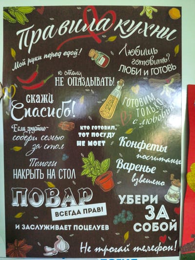 This is a photograph of a colorful, hand-painted poster on a dark brown background, likely displayed in a kitchen or a culinary-themed space. The poster features various handwritten phrases and illustrations related to cooking and family.
The dominant text in large, stylized white lettering at the top reads "Правила кухни" (Kitchen Rules). Below this, smaller text in white includes phrases like "Мой руки перед едой!" (Wash your hands before eating!), "к столу НЕ ОПАЗДЫВАТЬ!" (Don't be late for the table!), and "скажи Спасибо!" (Say Thank You!).
Illustrations peppered across the poster include red chili peppers, a bottle of oil, a jar labeled "PAPRIKA," cinnamon sticks, vanilla beans, a potted plant with green leaves, blueberries, garlic, and a spice grinder. There are also small hearts and leaves scattered throughout the design.
Further down, more text provides cooking-related advice and statements: "Любишь готовить? ЛЮБИ И ГОТОВЬ" (Love to cook? Love and cook), "готовим только с любовью" (We cook with love only), "Если грустно- собери семью за стол" (If you are sad, gather the family at the table), and "Помоги НАКРЫТЬ НА СТОЛ" (Help to set the table). There are also playful declarations like "ПОВАР ВСЕГДА ПРАВ!" (The chef is always right!) and "И ЗАСЛУЖИВАЕТ ПОЦЕЛУЕВ" (And deserves kisses).
Towards the bottom, a new set of rules is presented: "Конфеты посчитаны" (Candies are counted), "Варенье взвешено" (Jam is weighed), "УБЕРИ ЗА СОБОЙ" (Clean up after yourself), and a final strong directive, "Не трогай телефон!" (Don't touch your phone!).
The overall tone of the poster is warm, encouraging, and humorous, emphasizing the joy of cooking, family togetherness, and good kitchen etiquette. The vibrant colors and playful illustrations create a cheerful and inviting atmosphere. The poster appears to be in good condition, with no visible damage.