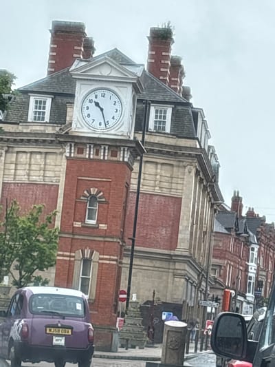 The image displays a prominent red-brick building with a distinctive clock tower, set in an urban environment in Trafford, United Kingdom. The sky is overcast, suggesting a cloudy day.
The building exhibits traditional architecture, characterized by its red brickwork, grey sloped roofs, and multiple tall brick chimneys. It features several windows with white frames; some are rectangular, while others are arched with decorative stone or concrete surrounds. The facade is adorned with horizontal bands of lighter-colored stone or concrete.
A white clock face, housed within a gabled structure on the building, indicates the time as approximately 4:25. The hour hand points between 4 and 5, and the minute hand points directly at 5.
In the foreground, partially visible on the right, is the rear of a purple car, possibly a taxi or private hire vehicle. Its license plate clearly reads "MJ58 OBA" and "W77". Adjacent to the car, parts of a street scene are visible, including a black lamppost and a red circular traffic sign. Green foliage from trees or bushes can be seen in the bottom right corner. The scene captures a quiet street on a cloudy afternoon.