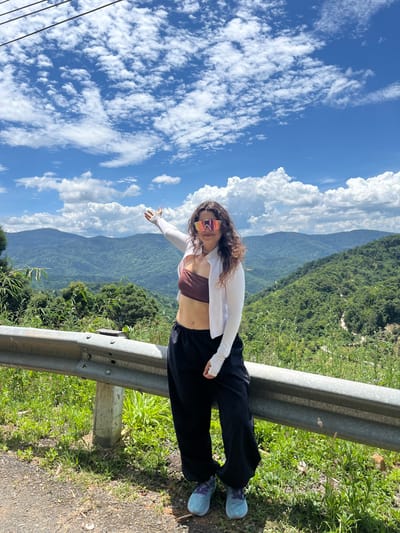 A woman stands on the side of a mountain road, her left arm extended upwards, gesturing towards a lush, green landscape. She is wearing a brown bandeau top, a white zip-up jacket, black loose-fitting pants, and blue sneakers. Her face is obscured by large, reflective sunglasses with pink and orange lenses. The road is bordered by a metal guardrail and overgrown grass. In the background, rolling hills covered in dense vegetation stretch towards a bright blue sky filled with fluffy white clouds. The scene appears to be in Son Dien, Vietnam, given the distinctive mountainous terrain and vegetation.