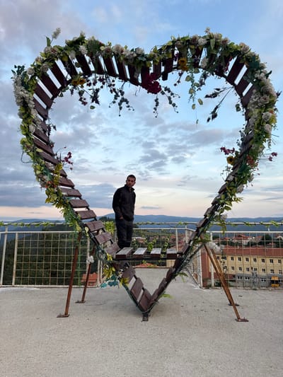 In the city of Sveta Petka, Bulgaria, a man stands within a large, heart-shaped wooden structure adorned with artificial flowers and greenery. The structure is positioned on a rooftop or elevated platform with a concrete floor. In the background, rolling hills and a residential area with buildings are visible under a cloudy sky. A metal railing with a wire mesh fence runs along the edge of the platform, separating it from the landscape below. The man is wearing a dark jacket and dark pants and is looking away from the camera. The heart structure has wooden slats that form a staircase-like design.