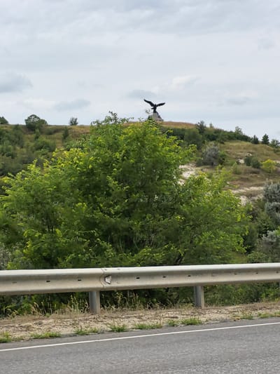 A dark, silhouetted statue of an eagle with outstretched wings stands atop a grassy hill under a cloudy sky. In the foreground, a metal guardrail separates a road from the landscape. Lush green trees and scrubby vegetation cover the lower slopes of the hill. The image appears to be taken from a roadside vantage point, possibly looking towards a monument in Belogorskiy, Russia.