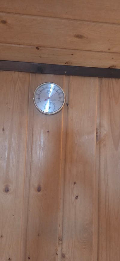 The image shows a close-up view of a round, analog thermometer and hygrometer mounted on a wooden wall. The instrument has a silver-colored bezel and a white face with markings for temperature in Fahrenheit, ranging from 10 to 120 degrees. A red needle indicates the temperature, pointing to approximately 80 degrees Fahrenheit. Below the main thermometer, there is a smaller dial for humidity, with markings from 0 to 100 percent. A blue needle indicates the humidity level, pointing to around 20 percent. The instrument is labeled "THERMO-HYGROMETER" and "SAUNA." The wooden wall appears to be made of vertical planks with visible knots and wood grain. A dark horizontal trim piece is present above the thermometer. The scene is indoors, likely in a sauna due to the instrument's labeling. There are no people or animals visible in the image. The location is unknown, and cannot be determined from the image itself.