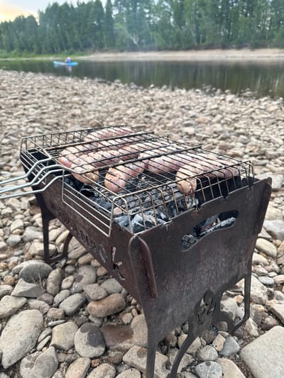 An outdoor scene unfolds on a rocky riverbank, with a focus on a portable metal grill filled with uncooked sausages. The grill, weathered and dark, sits on a bed of grey and tan pebbles that extend to the water's edge. Behind the grill, the calm, dark water of a river reflects a hazy, green treeline. In the distance, a small, blue inflatable raft with a person in a red shirt is visible on the water. The sky above is overcast, with a soft, diffused light. The overall impression is of a serene, natural setting, likely in a rural area, possibly during an outdoor recreational activity. Given the location context of Первомайское, Russia, this image likely depicts a moment of leisure by a local river.