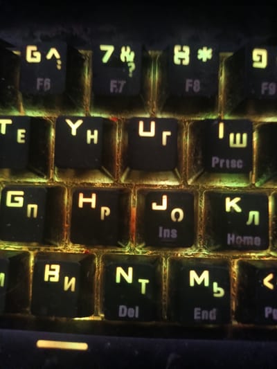 The image shows a close-up of a backlit keyboard, displaying a mix of Cyrillic and Latin characters, with some keys also showing function labels like "F6", "F7", "F8", "F9", "Prisc", "Ins", and "Home". The keyboard appears to be used and has some visible dust and debris. The lighting suggests a dimly lit environment. The keyboard's design and the presence of Cyrillic characters are consistent with a keyboard used in Russia. Specifically, the location of Шипуново, Russia, is mentioned in the prompt. This suggests the keyboard is situated within that geographic region.