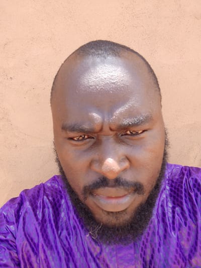 A close-up shot shows a man with dark skin, a bald head, and a full black beard wearing a patterned purple shirt. He has a serious expression with furrowed brows. The background appears to be a plain, textured wall of a light brown color. There is no information in the image to confirm the location is Mulke Barikin Sayejo, Nigeria.