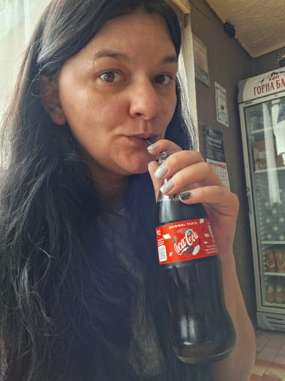 This media file shows a close-up, low-angle shot of a woman lying down and drinking from a bottle of Coca-Cola. Her long, dark hair covers a significant portion of the frame, obscuring much of her face and body. The woman's eye is visible, looking directly at the camera with a neutral expression. She is holding the Coca-Cola bottle with her right hand, and her fingernails are painted with a glittery silver and a dark gray polish. The bottle itself features the iconic red Coca-Cola label with white lettering.

In the background, there is a glimpse of a refrigerated display case filled with drinks and a calendar affixed to a wall. Text on the calendar is partially visible, along with the word "TRIO DENT" and what appears to be Bulgarian text, "Марин & Марини". Further text on the display case reads "ГОРНА БА" which likely is part of a brand name. The lighting suggests it is daytime, possibly indoors. The overall scene appears to be casual and relaxed, with the woman enjoying a beverage. The context of Marchevo, Bulgaria, indicates a potential Eastern European setting.