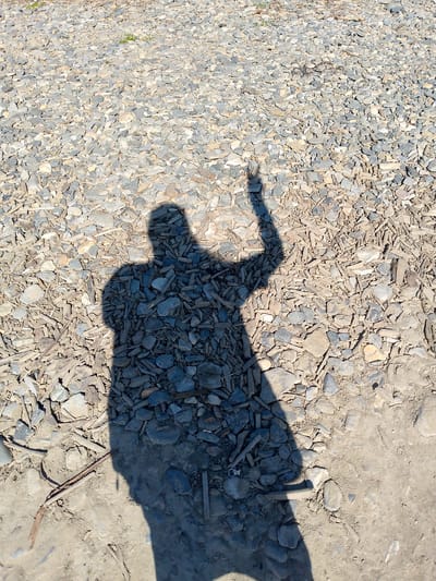 The image is an aerial shot of a shadow of a person standing on a surface covered with small gray and beige stones, with some dirt and small pieces of wood scattered throughout. The shadow is cast by the sun and appears to be in the shape of a person with their right arm raised. The person's shadow is dark and covers a significant portion of the image. The overall scene suggests an outdoor environment, possibly a path or a clearing, with natural elements. The image does not provide any specific visual cues to determine its location within the city of Magadan, Russia.