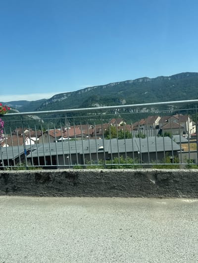 The image captures a view of a town nestled amidst rolling green hills and a distant mountain range under a clear blue sky. In the foreground, a grey metal fence runs horizontally across the frame, separating the viewer from a cluster of buildings with red-tiled roofs. Some of these buildings appear to be residential homes, while others might be part of a commercial area. Green foliage and trees are visible between the buildings, suggesting a well-maintained environment. The overall scene depicts a tranquil, sun-drenched day in a rural or semi-rural setting, likely in the French Jura region given the mention of Lavans-lès-Saint-Claude. The lack of visible people suggests a quiet moment, perhaps during the mid-day.