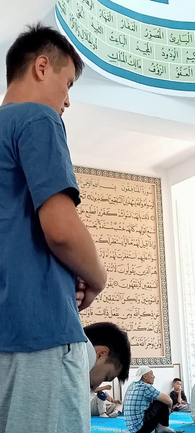 In a mosque in Besagash, Kazakhstan, a group of people are seen engaged in prayer. The primary focus is on a man in the foreground, seen from the back, wearing a blue t-shirt and light grey pants. He is standing and looking downwards. To his left, another man is partially visible, also looking down, wearing a blue and white checkered shirt and dark pants. In the background, a young boy in a black shirt and light pants is seated, looking towards the right. Another man, wearing a white cap and a blue and white checkered shirt, is seated to the right of the first man, also looking to the right. The walls of the mosque are decorated with Islamic calligraphy. The floor is covered with a blue carpet. The scene suggests a prayer service is in progress.